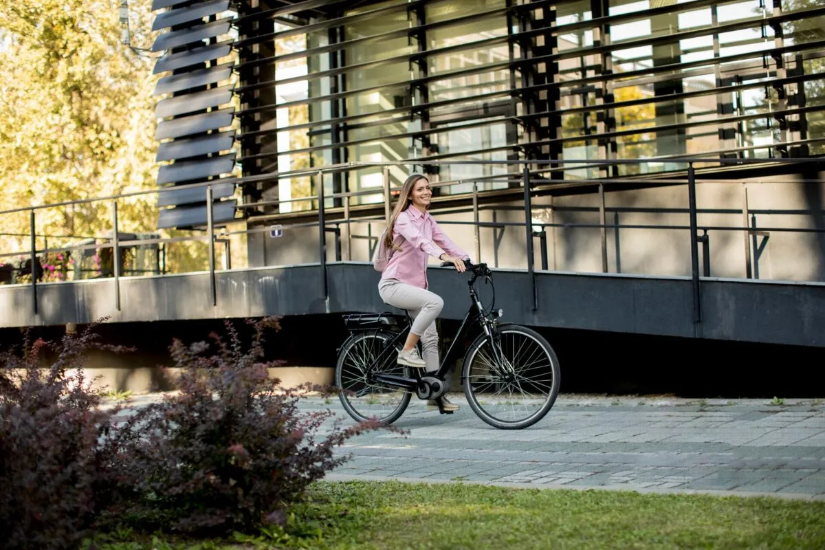 Uma mulher com camisa rosa pedalando uma bicicleta elétrica em um cenário urbano moderno.